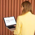 Weronika Wolska – ekspertka od projektowania graficznego – przy komputerze podczas pracy nad identyfikacją wizualną. W żółtej marynarce, odwrócona tyłem, w trakcie wyboru fontu do projektu graficznego. Grafika komputerowa, design, edukacja graficzna, kursy online, marketing wizualny, Akademia Projektowania.