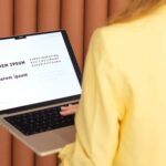 Weronika Wolska – założycielka Akademii Projektowania – podczas pracy nad projektem graficznym. W żółtej marynarce, odwrócona tyłem, przy komputerze w trakcie poszukiwania idealnego fontu. Nowoczesne brązowe tło, atmosfera skupienia i kreatywnego procesu. Grafika, design, projektowanie graficzne, reklama, marketing, edukacja wizualna.