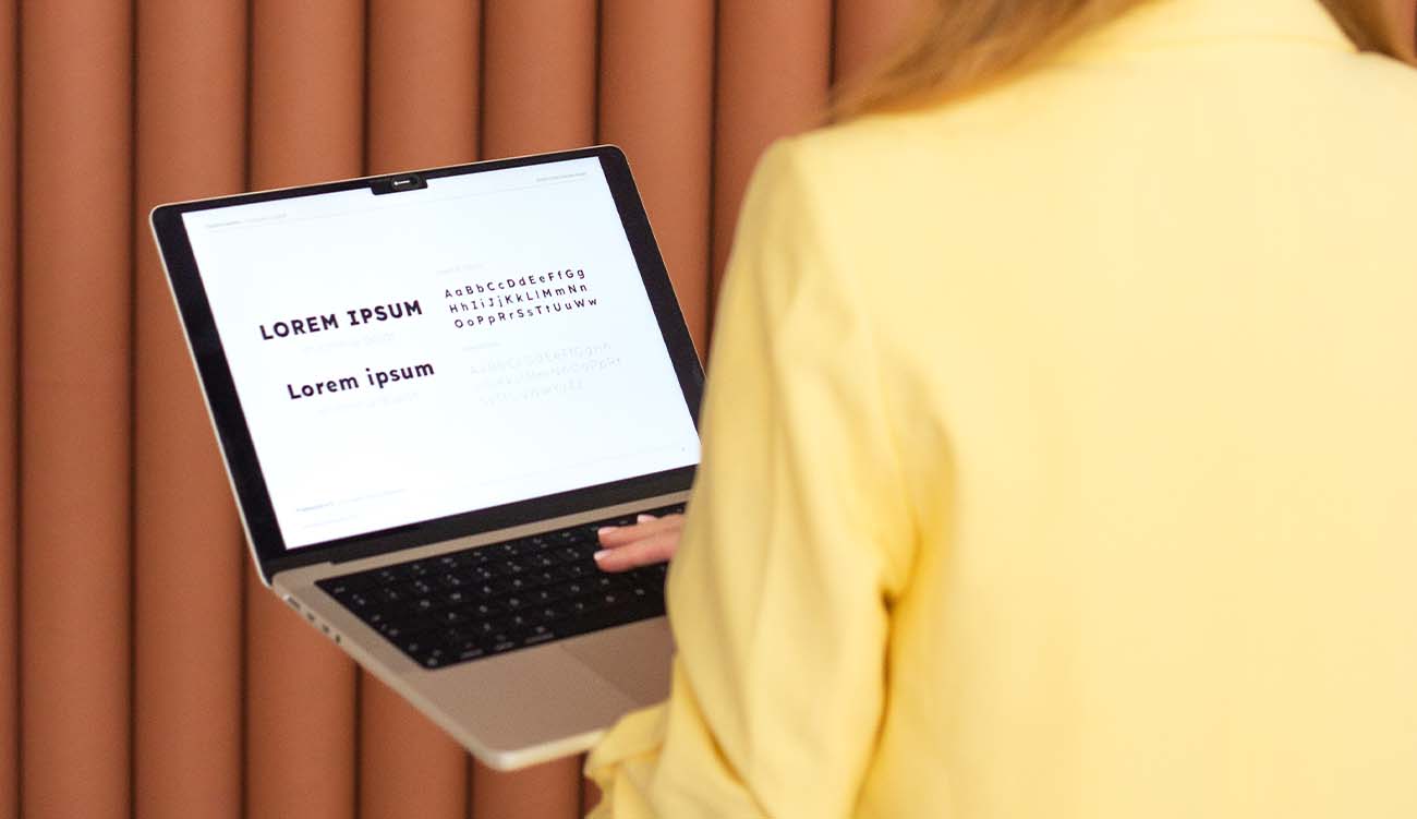 Weronika Wolska – założycielka Akademii Projektowania – podczas pracy nad projektem graficznym. W żółtej marynarce, odwrócona tyłem, przy komputerze w trakcie poszukiwania idealnego fontu. Nowoczesne brązowe tło, atmosfera skupienia i kreatywnego procesu. Grafika, design, projektowanie graficzne, reklama, marketing, edukacja wizualna.