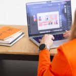 Weronika Wolska – twórczyni Akademii Projektowania – pracuje przy laptopie nad projektami graficznymi w programie do edycji. W tle widoczne są szkicowniki i książki o designie. Zdjęcie ilustruje artykuł blogowy z wywiadem z Aleksandrą Chlebowską – absolwentką Kursu Projektowania Graficznego, która dzieli się swoją ścieżką nauki grafiki komputerowej, budowy portfolio i pracy w branży kreatywnej jako początkująca freelancerka.