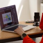 Weronika Wolska – projektantka graficzna i mentorka – siedzi przy biurku i pracuje na laptopie w Adobe Illustrator. Na ekranie widoczny projekt identyfikacji wizualnej. Obok mikrofon, notesy i palety kolorów. Zdjęcie przedstawia autorkę Kursu Projektowania Graficznego w Akademii Projektowania podczas pracy twórczej. To ujęcie osoby aktywnej w branży designu, grafiki komputerowej i edukacji graficznej, inspirującej innych do działania na własnych zasadach jako freelancer lub grafik kreatywny.