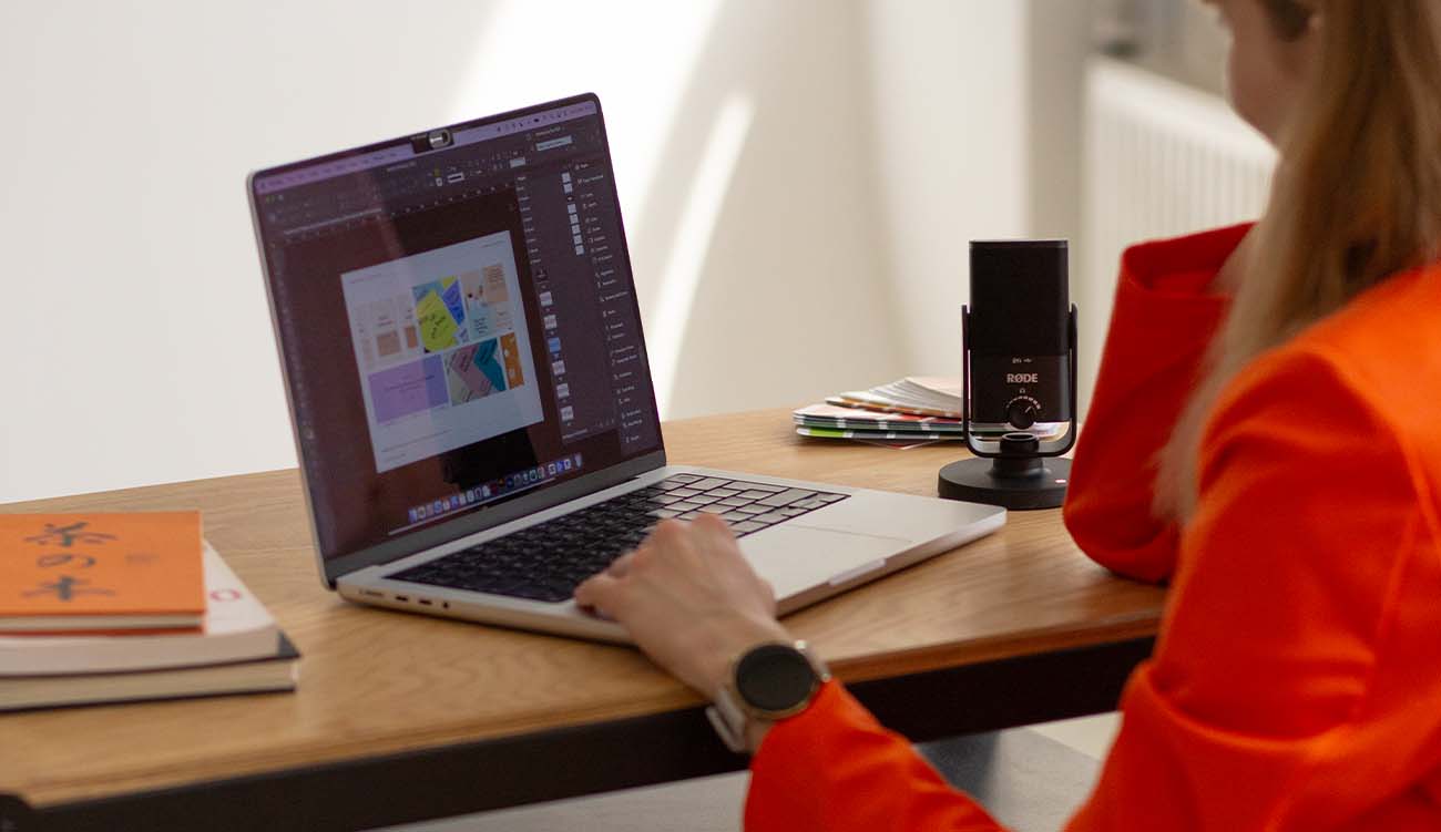 Weronika Wolska – projektantka graficzna i mentorka – siedzi przy biurku i pracuje na laptopie w Adobe Illustrator. Na ekranie widoczny projekt identyfikacji wizualnej. Obok mikrofon, notesy i palety kolorów. Zdjęcie przedstawia autorkę Kursu Projektowania Graficznego w Akademii Projektowania podczas pracy twórczej. To ujęcie osoby aktywnej w branży designu, grafiki komputerowej i edukacji graficznej, inspirującej innych do działania na własnych zasadach jako freelancer lub grafik kreatywny.