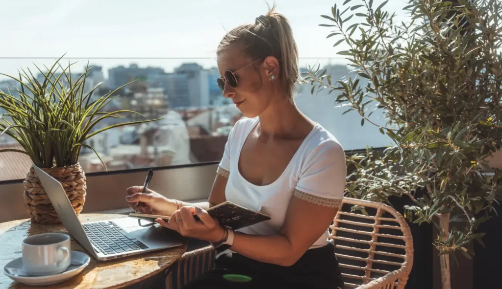 Klaudia Pawelec – absolwentka Kursu Projektowania Graficznego w Akademii Projektowania Weroniki Wolskiej – pracuje jako freelancerka nad projektem graficznym na tarasie, korzystając z laptopa i szkicownika. Obraz ukazuje mobilną pracę grafika komputerowego, projektowanie brandingowe oraz kreatywne podejście do pracy w branży designu i identyfikacji wizualnej.
