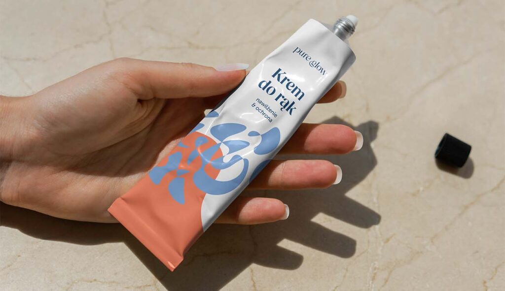 Początki w branży graficznej. Zdjęcie przedstawia rękę trzymającą minimalistyczną tubkę kremu do rąk marki Pure Glow. Opakowanie utrzymane jest w estetycznej, nowoczesnej kolorystyce – biel, pastelowy niebieski i pomarańczowy. Etykieta zawiera nazwę produktu „Krem do rąk – nawilżenie & ochrona” oraz ozdobną, abstrakcyjną ilustrację wektorową. Projekt graficzny opakowania został stworzony przez Karolinę Krajewską, absolwentkę Kursu Projektowania Graficznego w Akademii Projektowania, i prezentowany jest w artykule Weroniki Wolskiej jako przykład współczesnego brand designu i grafiki użytkowej.
