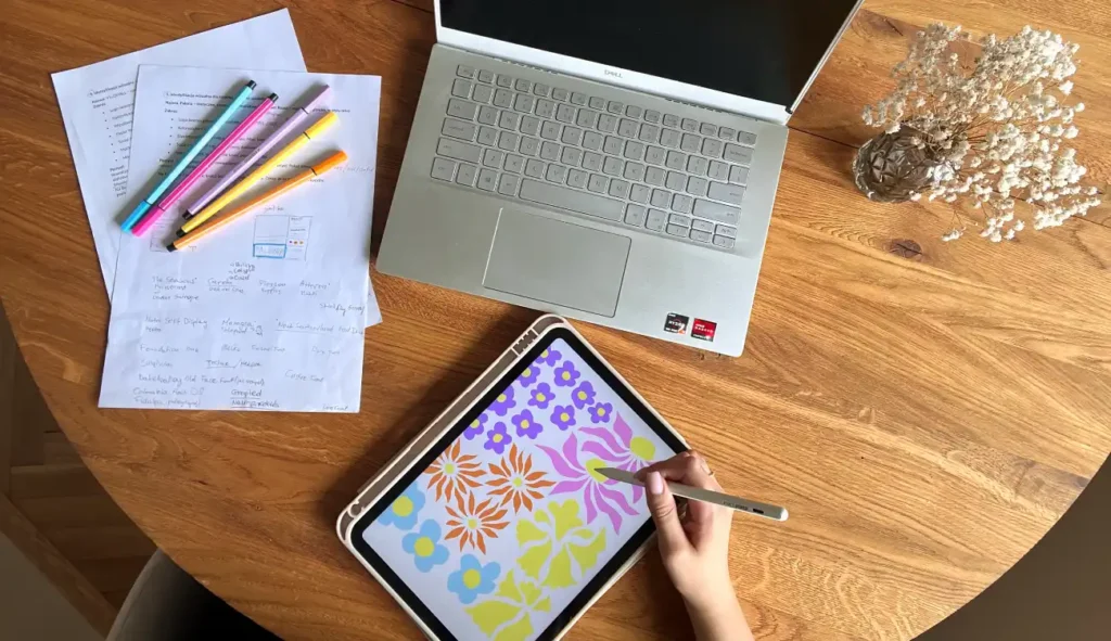Zdjęcie przedstawia miejsce pracy Aleksandry Chlebowskiej – absolwentki Kursu Projektowania Graficznego w Akademii Projektowania Weroniki Wolskiej. W kadrze widoczny jest tablet z rysowaną cyfrową ilustracją kwiatową, laptop, kolorowe flamastry, notatki projektowe oraz szkice koncepcyjne. To ujęcie z procesu twórczego młodej graficzki komputerowej pracującej zdalnie nad projektami graficznymi i rozwijającej swoje umiejętności w zakresie ilustracji, projektowania i designu w ramach kursu online.