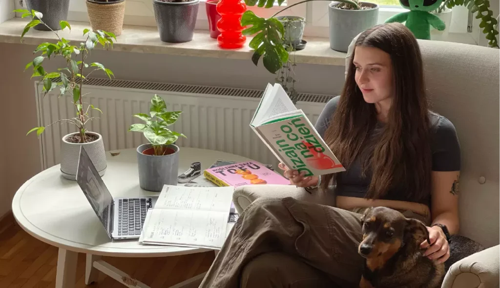 Sara Zimorodzka, absolwentka Kursu Projektowania Graficznego w Akademii Projektowania prowadzonej przez Weronikę Wolską, siedzi w fotelu z psem na kolanach w swoim domowym miejscu pracy. Na stole obok znajduje się laptop z otwartym projektem graficznym w programie do edycji grafiki komputerowej, notatki, książka „Dizajn na co dzień” i magazyny o designie. W tle rośliny doniczkowe nadają wnętrzu przytulny klimat. Scena ukazuje twórczą codzienność freelancera i grafika – pracę nad projektowaniem graficznym, designem, UX/UI, brandingiem i ilustracją cyfrową w przestrzeni sprzyjającej kreatywności.
